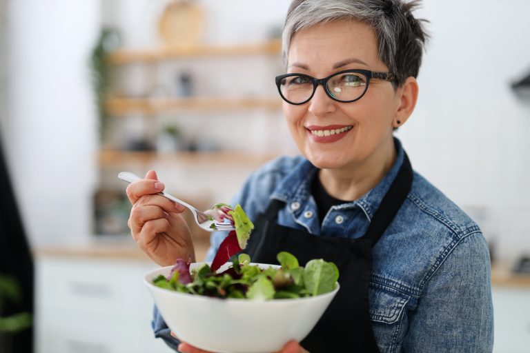 Portrait of a smiling mature woman in glasses with a large plate of salad in the kitchen